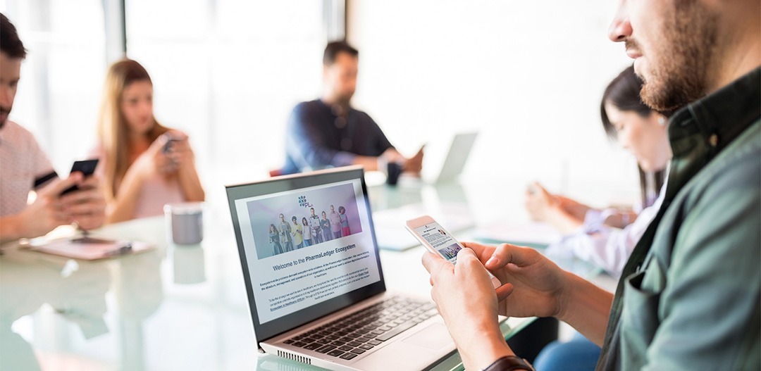 Man with both his phone and computer searching the PharmaLedger website for blockchain solutions for healthcare during a meeting break with his coworkers in the background.